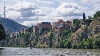 قارب سياحي يبحر في نهر كورا بجانب المدينة القديمة في تبليسي مع إطلالة على قلعة ناريكالا والصخور الجبلية